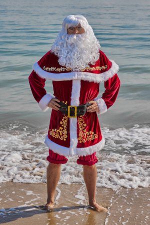 Téléchargez les photos : Père Noël en mer par une chaude journée d'été. Debout, la cheville dans l'eau, le pantalon enroulé. Vacances de Noël ou du Nouvel An dans les pays chauds. Vacances d'été du Père Noël. - en image libre de droit