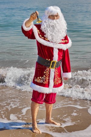 Téléchargez les photos : Père Noël en mer par une chaude journée d'été. Père Noël se tient cheville profonde dans l'eau avec son pantalon enroulé et sonne une cloche. Vacances de Noël ou du Nouvel An dans les pays chauds. Vacances d'été du Père Noël. - en image libre de droit