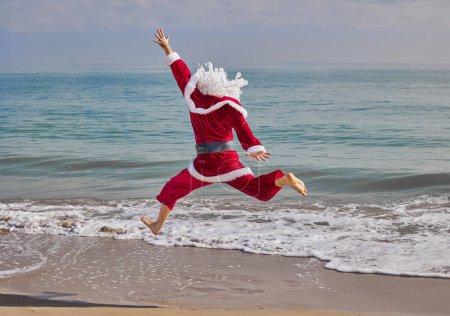Téléchargez les photos : Père Noël en mer par une chaude journée d'été. Père Noël, vu de derrière, sautant sur la plage face à la mer. Vacances de Noël ou du Nouvel An dans les pays chauds. Vacances d'été du Père Noël. - en image libre de droit