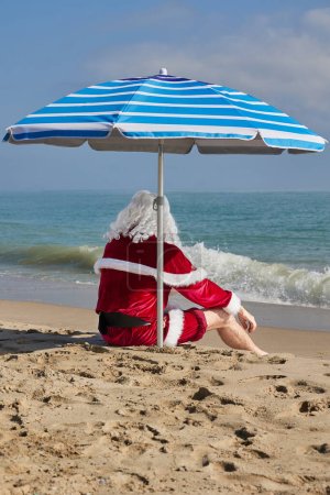 Téléchargez les photos : Père Noël en mer par une chaude journée d'été. Vue arrière du Père Noël assis sous un parapluie sur la plage donnant sur la mer. Vacances de Noël ou du Nouvel An dans les pays chauds. Vacances d'été du Père Noël. - en image libre de droit