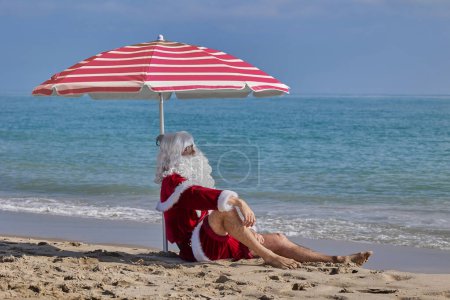 Téléchargez les photos : Père Noël en mer par une chaude journée d'été. Père Noël assis sous un parapluie sur la plage en face de la mer. Vacances de Noël ou du Nouvel An dans les pays chauds. Vacances d'été du Père Noël. - en image libre de droit