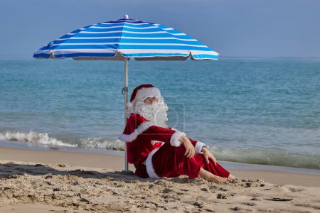 Téléchargez les photos : Père Noël en mer par une chaude journée d'été. Père Noël assis sous un parapluie sur la plage en face de la mer. Vacances de Noël ou du Nouvel An dans les pays chauds. Vacances d'été du Père Noël. - en image libre de droit