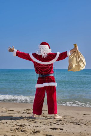 Téléchargez les photos : Père Noël en mer par une chaude journée d'été. Vue arrière du Père Noël debout pieds nus les bras tendus sur une plage de sable en face de la mer. Il tient un sac avec des cadeaux à la main.. - en image libre de droit