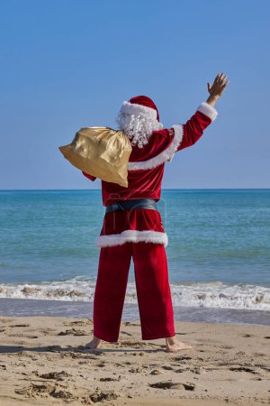 Téléchargez les photos : Père Noël en mer par une chaude journée d'été. Vue arrière du Père Noël debout pieds nus sur une plage de sable en face de la mer. Il tient un sac avec des cadeaux sur l'épaule et agite la main.. - en image libre de droit