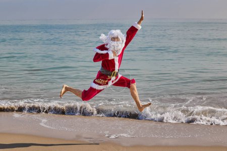 Téléchargez les photos : Le Père Noël saute sur la plage dans le contexte de la mer par une chaude journée d'été. Vacances de Noël ou du Nouvel An dans les pays chauds. Vacances d'été du Père Noël. - en image libre de droit