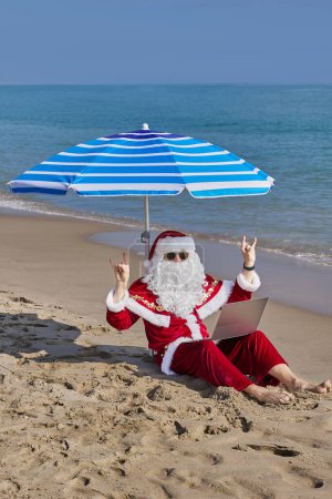 Téléchargez les photos : Le Père Noël est assis sous un parasol sur le fond de la mer par une chaude journée d'été et tape quelque chose sur un ordinateur portable. Vacances de Noël ou du Nouvel An dans les pays chauds. Vacances d'été du Père Noël. - en image libre de droit