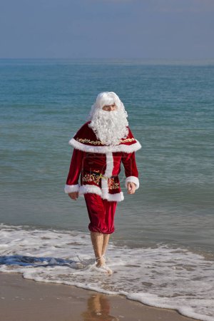 Téléchargez les photos : Le Père Noël se promène le long de la ligne de surf sur la plage par une chaude journée d'été. Vacances de Noël ou du Nouvel An dans les pays chauds. Vacances d'été du Père Noël. - en image libre de droit