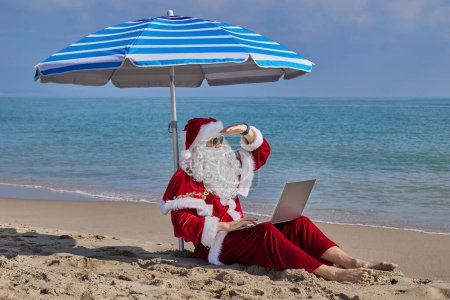 Téléchargez les photos : Le Père Noël est assis sous un parasol sur le fond de la mer par une chaude journée d'été et tape quelque chose sur un ordinateur portable. Vacances de Noël ou du Nouvel An dans les pays chauds. Vacances d'été du Père Noël. - en image libre de droit