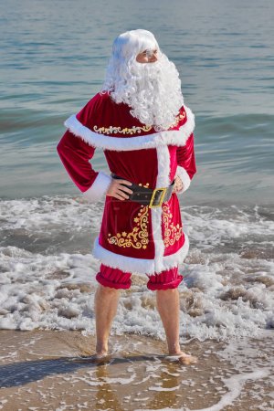 Téléchargez les photos : Père Noël en mer par une chaude journée d'été. Debout, la cheville dans l'eau, le pantalon enroulé. Vacances de Noël ou du Nouvel An dans les pays chauds. Vacances d'été du Père Noël. - en image libre de droit