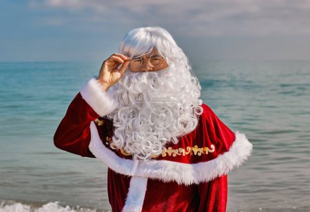 Téléchargez les photos : Gros plan portrait du Père Noël sur fond de mer par une chaude journée d'été. Le Père Noël ajuste ses lunettes. Vacances de Noël ou du Nouvel An dans les pays chauds. Vacances d'été du Père Noël. - en image libre de droit