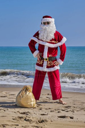 Téléchargez les photos : Père Noël en mer par une chaude journée d'été. Le Père Noël se tient pieds nus sur une plage de sable en face de la mer. Sac de cadeaux sur le sable. - en image libre de droit
