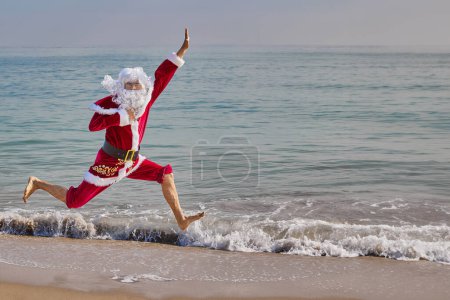 Photo for Santa Claus jumps on the beach against the backdrop of the sea on a hot summer day. Christmas or New Year holidays in hot countries. Santas summer vacation. - Royalty Free Image