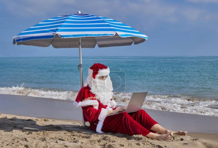 Téléchargez les photos : Le Père Noël est assis sous un parasol sur le fond de la mer par une chaude journée d'été et tape quelque chose sur un ordinateur portable. Vacances de Noël ou du Nouvel An dans les pays chauds. Santas vacances d'été. - en image libre de droit