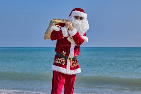 Téléchargez les photos : Père Noël en mer par une chaude journée d'été. Le Père Noël se tient pieds nus sur une plage de sable en face de la mer. Il tient un sac avec des cadeaux sur l'épaule et agite la main.. - en image libre de droit