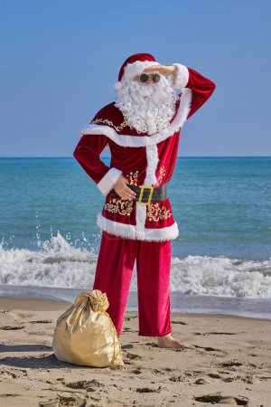 Téléchargez les photos : Père Noël en mer par une chaude journée d'été. Le Père Noël se tient pieds nus sur une plage de sable en face de la mer. Sac de cadeaux sur le sable. - en image libre de droit