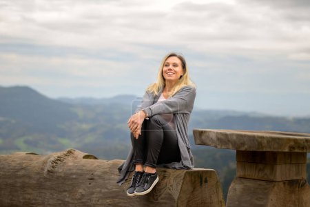 Téléchargez les photos : Joyeux rire femme blonde d'âge moyen assis sur un tronc d'arbre sur une montagne et ciel nuageux - en image libre de droit
