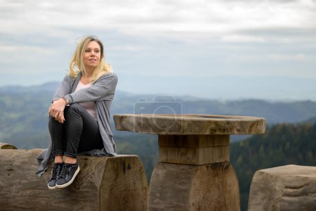 Téléchargez les photos : Heureuse jeune femme blonde d'âge moyen regardant au loin tout en étant assise sur une bûche au sommet d'une montagne et le ciel nuageux - en image libre de droit