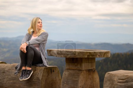 Téléchargez les photos : Heureuse jeune femme blonde d'âge moyen regardant au loin tout en étant assise sur une bûche au sommet d'une montagne et le ciel nuageux - en image libre de droit