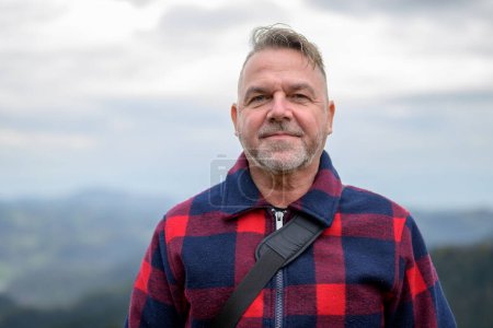 Foto de Hombre de pelo gris de mediana edad con una barba y una chaqueta de leñador está en una montaña con mal tiempo y mira a la cámara - Imagen libre de derechos