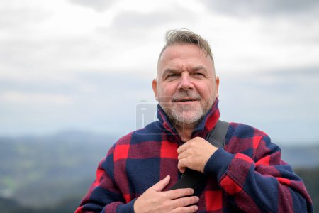 Foto de Hombre de pelo gris de mediana edad con una barba y una chaqueta de leñador está parado en una montaña con mal tiempo y está cerrando su chaqueta - Imagen libre de derechos