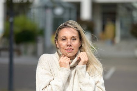Photo for Attractive blonde middle aged woman with wind tousled hair has her hands on her white turtleneck sweater stands against an urban backdrop with a gentle smile - Royalty Free Image