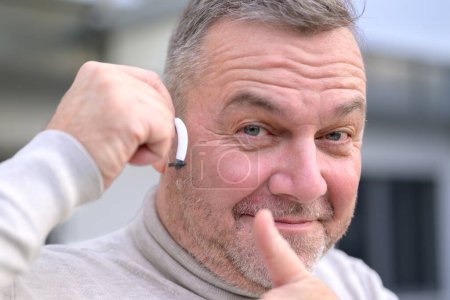 Téléchargez les photos : Portrait of a middle aged grey-haired hard of hearing man in his fifties or sixties with a hearing aid in his ear giving a thumbs up - en image libre de droit