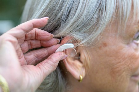 Téléchargez les photos : Older grey-haired deaf lady in her 80's with a hearing aid in her ear just putting it in - en image libre de droit