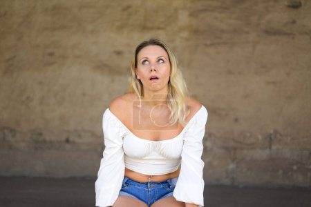 Téléchargez les photos : Portrait of a blond attractive woman with a white blouse with an annoyed look up and shows that she is annoyed in front of a brown wall - en image libre de droit