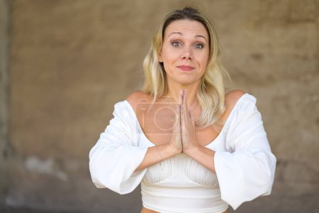 Foto de Portrait of a blond attractive woman with a white blouse with both hands folded so whether in church or pleading in front of a brown wall, - Imagen libre de derechos