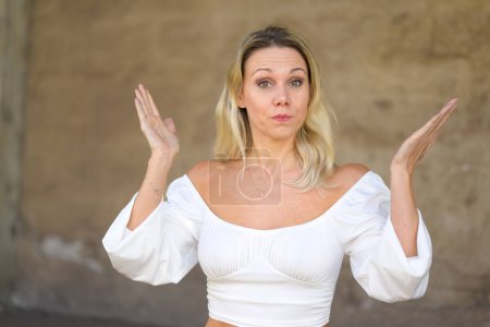 Foto de Portrait of a blond attractive woman with a white blouse with both hands to the side according to the motto I don't know or bad luck in front of a brown wall, outside - Imagen libre de derechos
