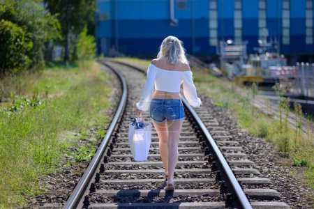 Téléchargez les photos : Back view of a blond woman with a white blouse and short jeans and a bag is walking on train tracks - en image libre de droit