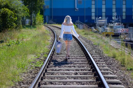 Foto de Vista trasera de una mujer rubia con una blusa blanca y pantalones vaqueros cortos y una bolsa está caminando en las vías del tren - Imagen libre de derechos