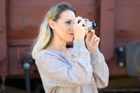 Téléchargez les photos : Vue latérale d'une femme d'âge moyen souriante avec un vieil appareil photo prend une photo et regarde à travers le viseur - en image libre de droit