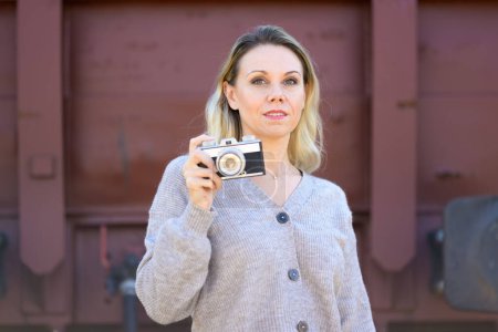 Photo for Front view of a serious middle aged woman holding an old camera wearing a brown sweater in front of an old train wagon - Royalty Free Image