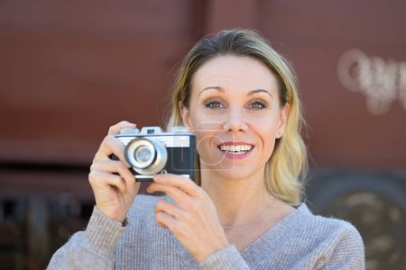 Téléchargez les photos : Vue de face d'une femme d'âge moyen souriante tenant une vieille caméra portant un pull marron devant un vieux wagon de train - en image libre de droit