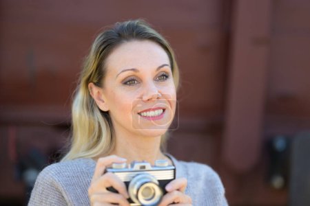 Photo for Side view of a smiling middle aged woman holding an old camera wearing a brown sweater in front of an old train wagon - Royalty Free Image