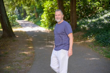Photo for Middle aged bearded man walking in a shady rural lane turning to smile at the camera - Royalty Free Image