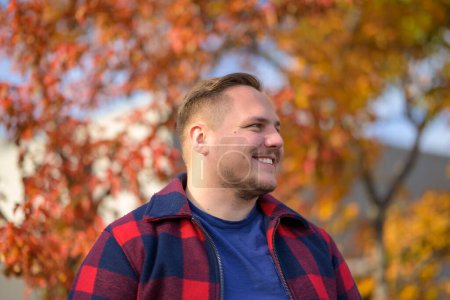 Photo for Young man with a bright smile in a peasant jacket stands in front of an autumn background with trees and yellow leaves - Royalty Free Image
