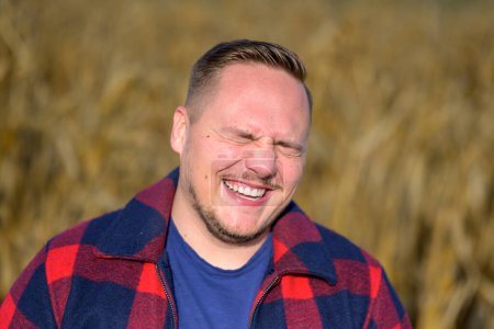 Photo for Portrait of a young man with a laughing fit of extreme joy with eyes closed wearing a red and blue lumberjack jacket and blue t-shirt against an autumnal cornfield as a background - Royalty Free Image