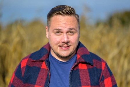 Foto de Joven con una chaqueta de leñador roja y azul y una camiseta azul frente a un campo de maíz de otoño mientras un fondo intenta mantener sus ojos abiertos contra la luz del sol. - Imagen libre de derechos