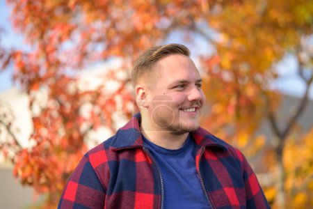 Téléchargez les photos : Vue latérale d'un jeune homme avec un sourire éclatant dans une veste paysanne se dresse devant un arrière-plan d'automne avec des arbres et des feuilles jaunes - en image libre de droit