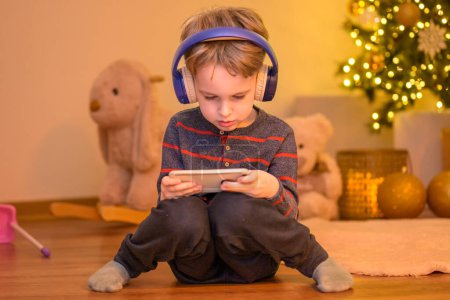 Foto de El niño de cinco años sentado en el piso de la sala de estar con auriculares y jugando con su teléfono móvil frente al árbol de Navidad decorado - Imagen libre de derechos