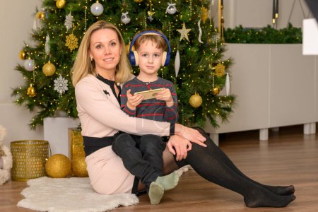 Téléchargez les photos : Le fils de cinq ans est assis sur les genoux de sa mère, joue avec son téléphone portable et la mère regarde la caméra devant le sapin de Noël décoré - en image libre de droit