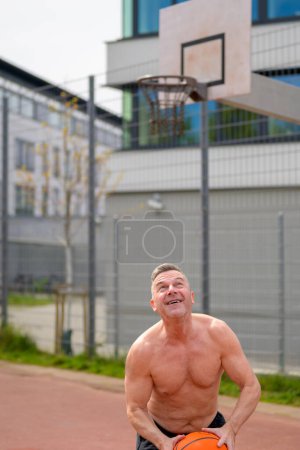 Foto de Hombre sin camisa de mediana edad en pantalones cortos deportivos negros a punto de saltar con el baloncesto en sus manos mirando concentrado a la cesta - Imagen libre de derechos