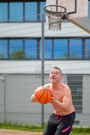 Foto de Hombre sin camisa de mediana edad en pantalones cortos deportivos negros a punto de saltar con el baloncesto en sus manos en un campo de deportes - Imagen libre de derechos