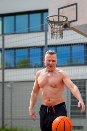 Téléchargez les photos : Homme torse nu du moyen âge en short de sport noir dribble et regarde vers le bas au basket sur un terrain de sport - en image libre de droit