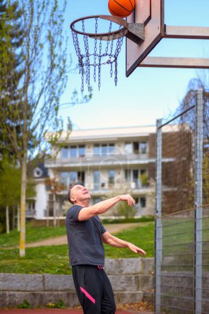 Foto de Hombre de mediana edad en pantalones cortos deportivos negros lanzando cestas mientras juega baloncesto en un campo de deportes - Imagen libre de derechos