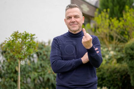 Foto de Hombre de mediana edad con el pelo gris, sin afeitar, con un suéter de cuello alto azul se encuentra en el jardín y muestra los dedos medios en la cámara - Imagen libre de derechos