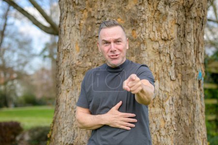 Téléchargez les photos : Homme aux cheveux gris debout devant un arbre dans le parc et pointant agressivement vers la caméra - en image libre de droit