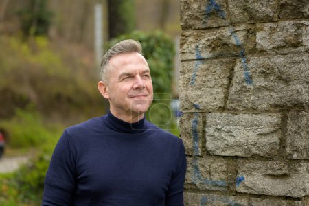 Photo for Middle age grey-haired attractive man wearing a blue sweater is standing near a wall of large stones and looking into the distance with a gentle smile - Royalty Free Image